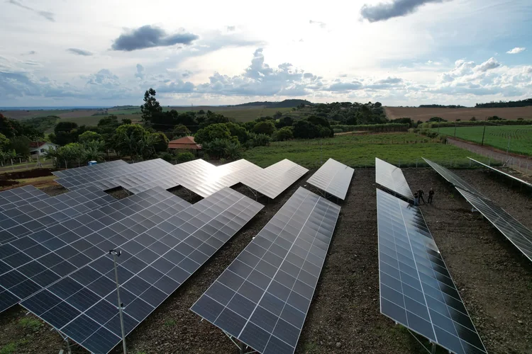 Vista aérea de usina fotovoltaica instalada no solo em propriedade rural, com fileiras de mesas metálicas e lavoura ao fundo